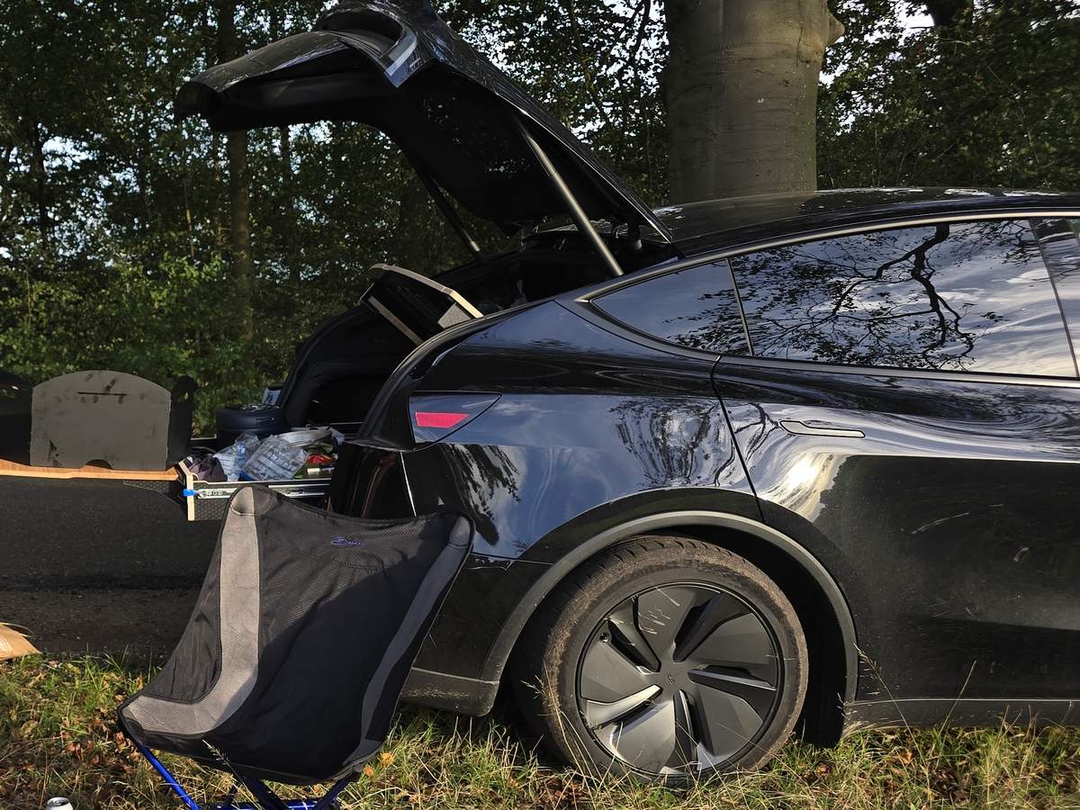 Zwarte Tesla Model Y met open achterklep in het gras aan een bosrand, met een opvouwbaar kampeerstoeltje ervoor