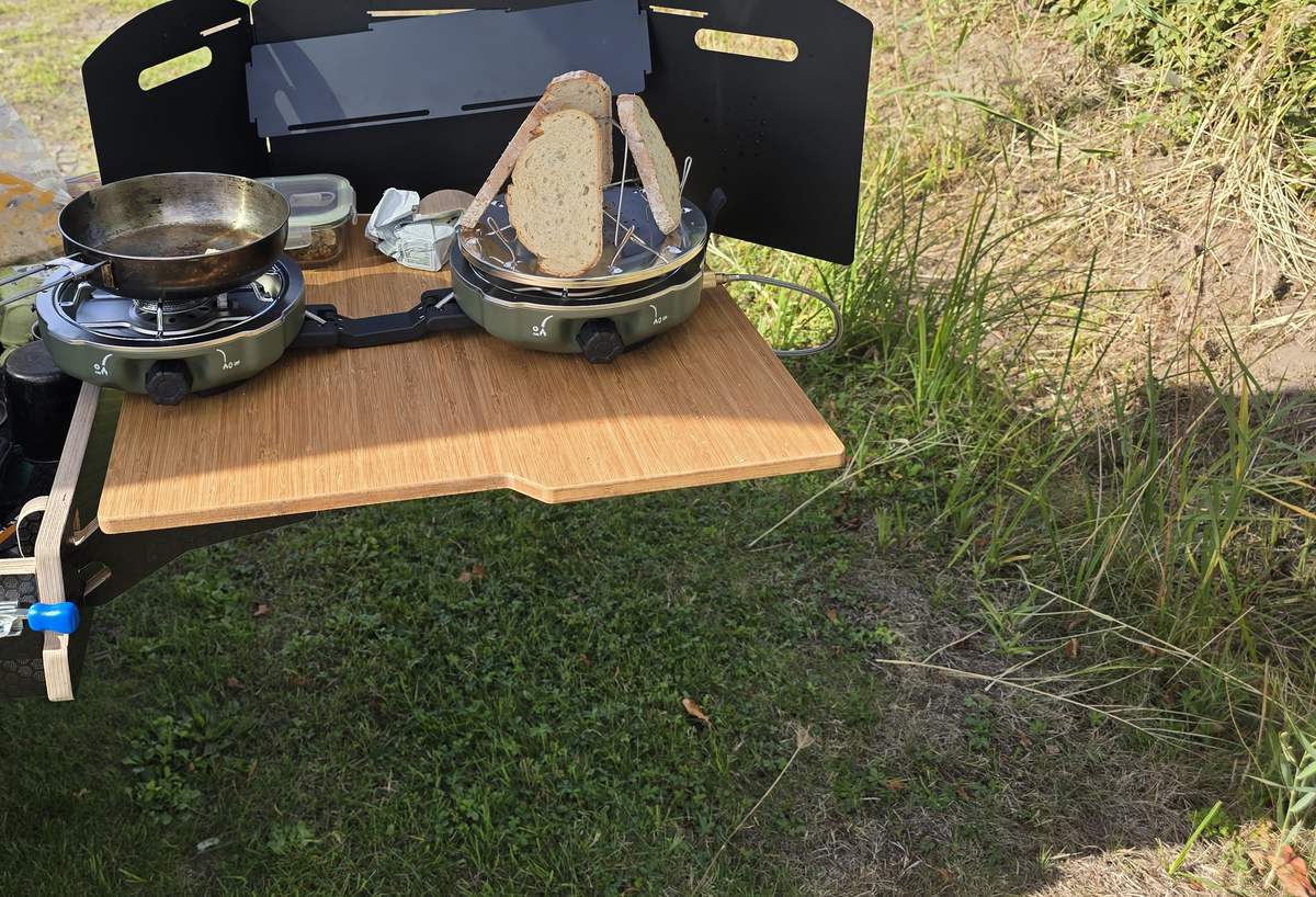Uitklapbare kampeerkeuken met twee gasbranders, een koekenpan en geroosterd brood in het gras